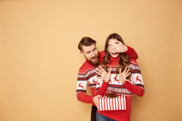 Guy closes the girls eyes with his hand and givs her a gift on a beige background in the studio.They both dressed in red and white sweaters with deer. - Фото, зображення