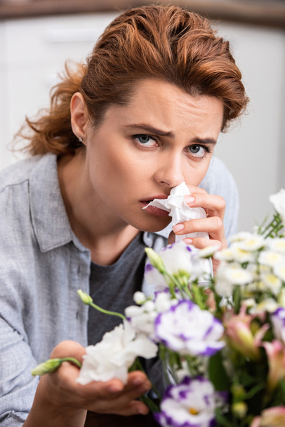 επιλεκτική εστίαση της άρρωστης γυναίκας με αλλεργία γύρη κρατώντας ιστό και αγγίζοντας τα λουλούδια  - Φωτογραφία, εικόνα