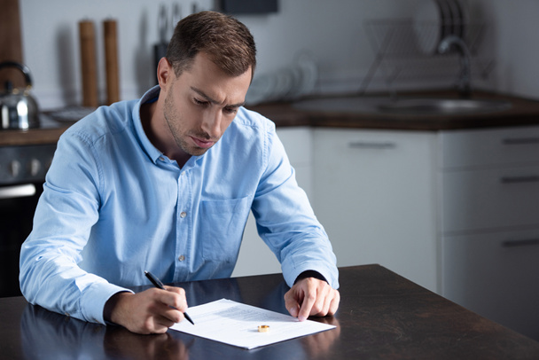 sad man in shirt sitting at table with ring and signing divorce documents - Fotografie, Obrázek
