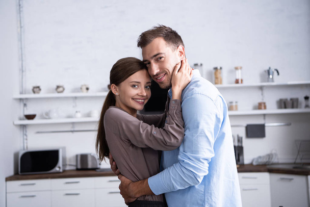 χαμογελαστός σύζυγος και ο σύζυγος αγκαλιάζοντας στην κουζίνα στο σπίτι - Φωτογραφία, εικόνα