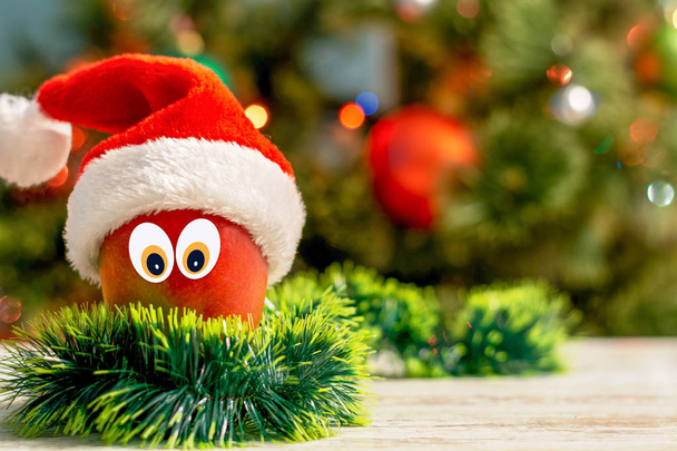 Año Nuevo y Navidad fondo conceptual con frutas tropicales. Mango con ojos en un sombrero de Papá Noel y un árbol de Navidad en el fondo. Vista frontal, espacio para texto, espacio para copiar
. - Foto, imagen