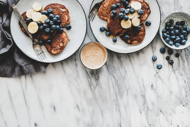 Healthy comfort breakfast set. Flat-lay of banana pancakes with fresh blueberry and honey and cup of coffee over white marble table background, top view, copy space - Фото, зображення