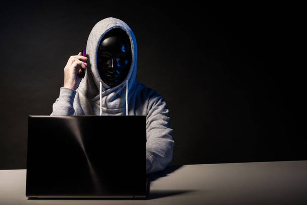 Ανώνυμος χάκερ στη μάσκα προγραμματιστής χρησιμοποιεί ένα φορητό υπολογιστή και μιλάει στο τηλέφωνο για να χαράξει το σύστημα στο σκοτάδι. - Φωτογραφία, εικόνα