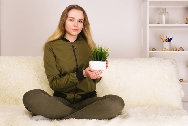 Молода дівчина-підліток з акне сидить на дивані і тримає зелену кімнатну рослину
 - Фото, зображення