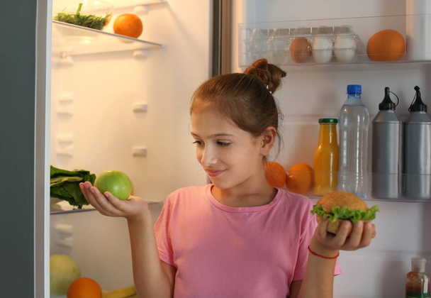 Девушка со здоровой и нездоровой пищей возле холодильника ночью
 - Фото, изображение