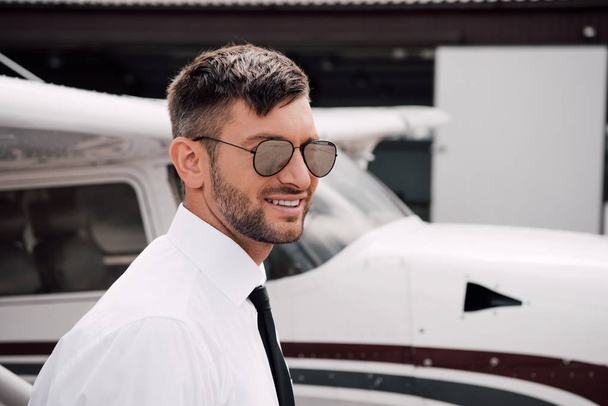 piloto barbudo en ropa formal y gafas de sol sonriendo cerca del avión
 - Foto, Imagen