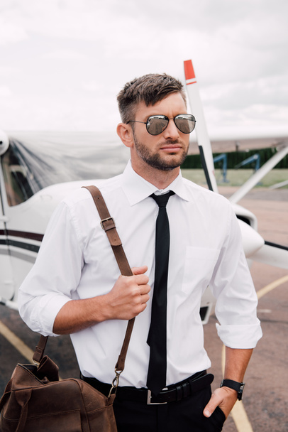 zelfverzekerd piloot in zonnebril met tas staan met de hand in zak in de buurt van het vliegtuig - Foto, afbeelding
