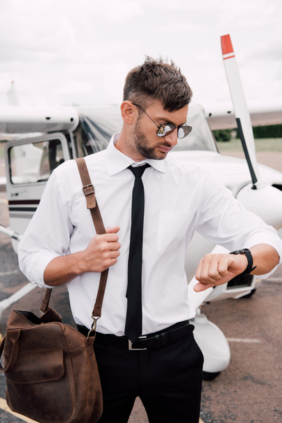 бородатый пилот в солнцезащитных очках стоит рядом с самолетом и смотрит на наручные часы
 - Фото, изображение