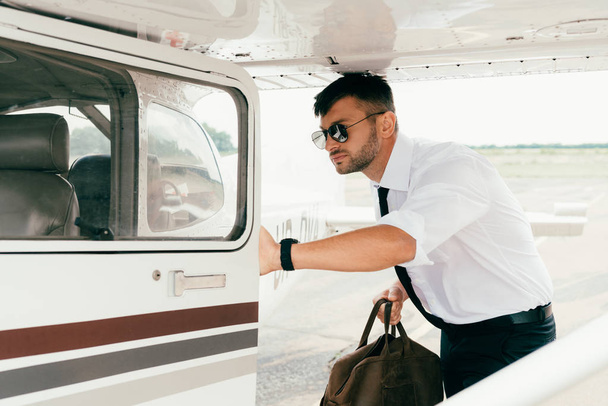 poważny pilot w okulary trzymając worek w pobliżu samolotu - Zdjęcie, obraz