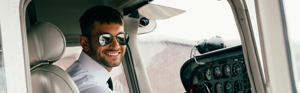 panoramic shot of smiling handsome pilot in sunglasses looking at camera in plane - Foto, Bild
