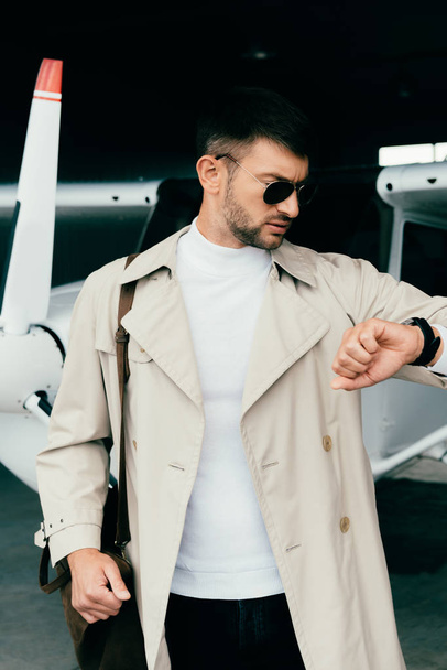 stylish businessman in coat standing near plane and looking at wristwatch - Фото, зображення