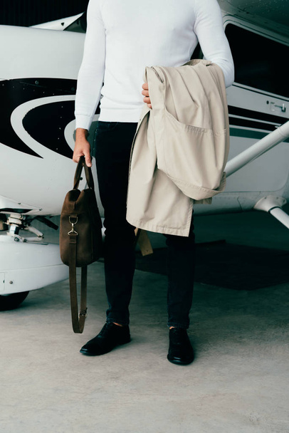 partial view of young man holding coat and bag while standing near plane - Foto, Imagen