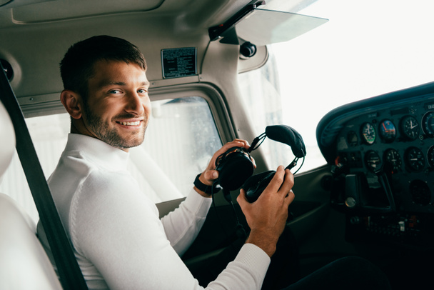 usměvavý vousatý mladý muž v příležitostných šatech držící sluchátka v letadle - Fotografie, Obrázek