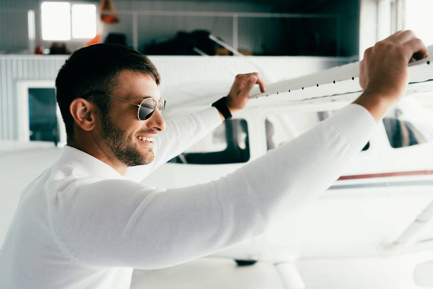вид сбоку улыбающегося бородатого мужчины в солнцезащитных очках, стоящего возле самолета
 - Фото, изображение