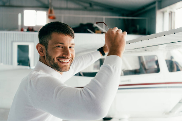smiling bearded man standing near plane and holding sunglasses - Foto, Imagem