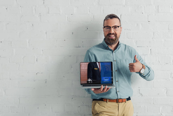handsome and smiling businessman showing thumb up and holding laptop with booking website  - Fotoğraf, Görsel