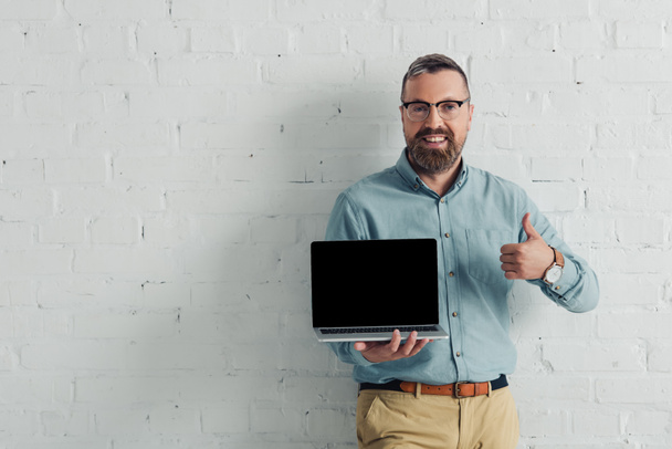 handsome and smiling businessman showing thumb up and holding laptop with copy space  - Zdjęcie, obraz