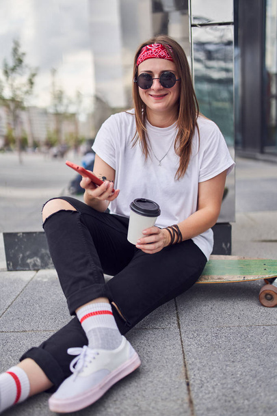 Photo of brunette in sunglasses with phone and glass in her hands sitting on skateboard on background of modern buildings in city - Фото, изображение