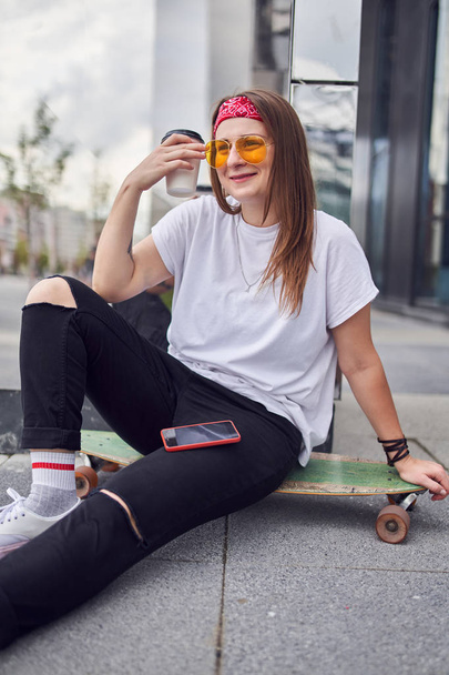 Photo of young brunette in sunglasses with glass in her hands sitting on skateboard on background of modern buildings in city - Φωτογραφία, εικόνα