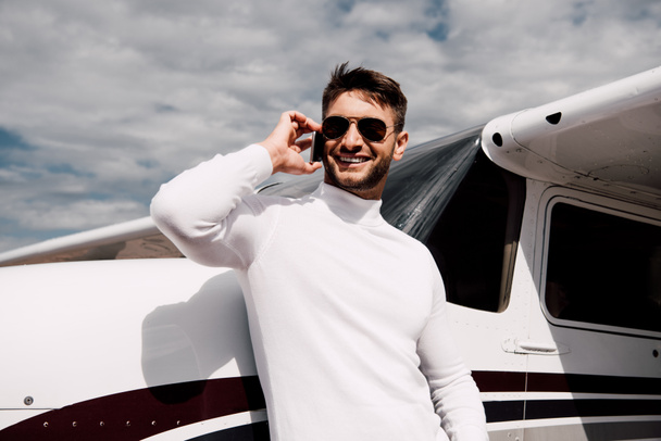 smiling bearded man in sunglasses talking on smartphone near plane - Foto, afbeelding