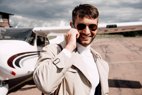 smiling man in coat with bag talking on smartphone near plane - Фото, зображення