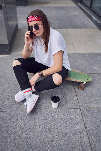 Photo of young brunette in sunglasses talking on phone sitting on skateboard on background of modern buildings in city - Fotografie, Obrázek