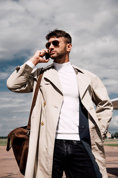 low angle view of pensive man in coat with bag talking on smartphone near plane - Zdjęcie, obraz