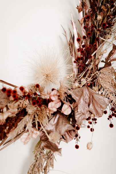 Vertical closeup shot of round twig wreath with leaves and red flowers on a white wall - 写真・画像
