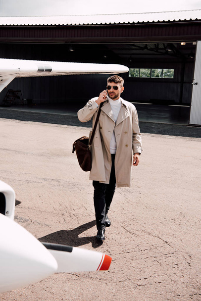 full length view of man in coat with bag talking on smartphone near plane in sunny day - 写真・画像