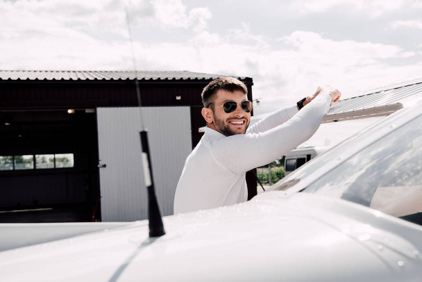 homem barbudo sorridente em óculos de sol de pé perto do avião em dia ensolarado
 - Foto, Imagem