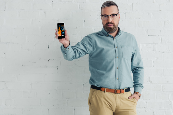 красивий бізнесмен з рукою в кишені тримає смартфон з графіками і сайтом графіків
  - Фото, зображення