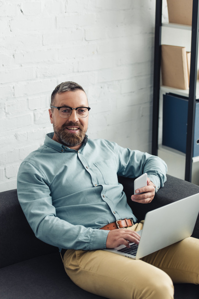 όμορφος επιχειρηματίας σε πουκάμισο κρατώντας φορητό υπολογιστή και smartphone στο γραφείο  - Φωτογραφία, εικόνα