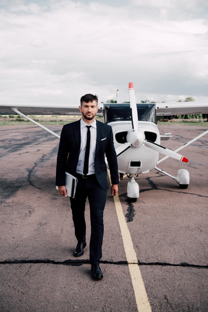 full length view of businessman in formal wear holding laptop near plane in sunny day - Photo, Image