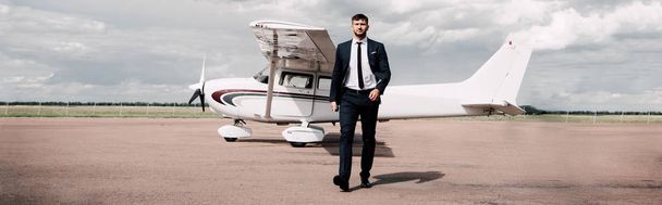 panoramic shot of businessman in formal wear walking near plane in sunny day - Photo, Image