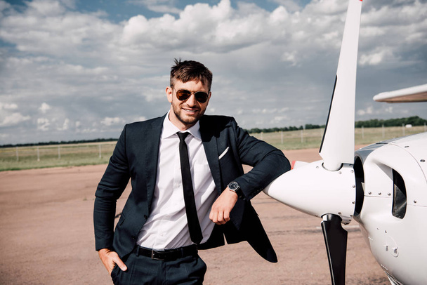 smiling businessman in formal wear standing with hand in pocket near plane - Zdjęcie, obraz