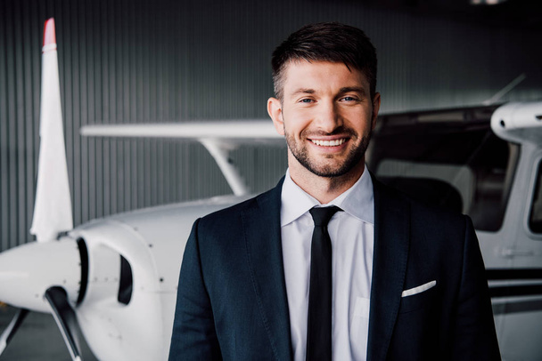vista frontal de homem de negócios sorridente em desgaste formal de pé perto do avião e olhando para a câmera
 - Foto, Imagem