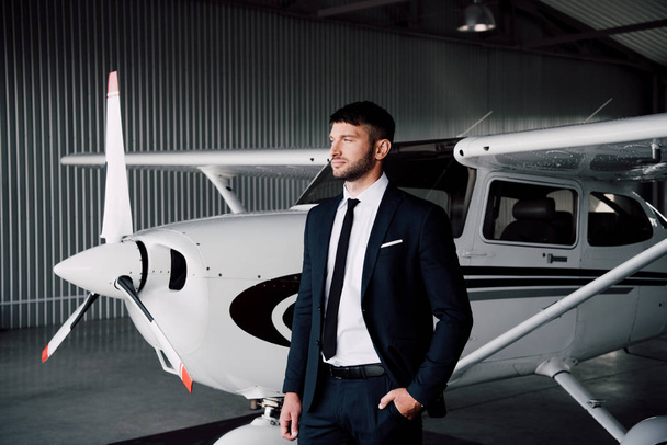 zelfverzekerde zakenman in formele slijtage staande met de hand in zak in de buurt van het vliegtuig - Foto, afbeelding