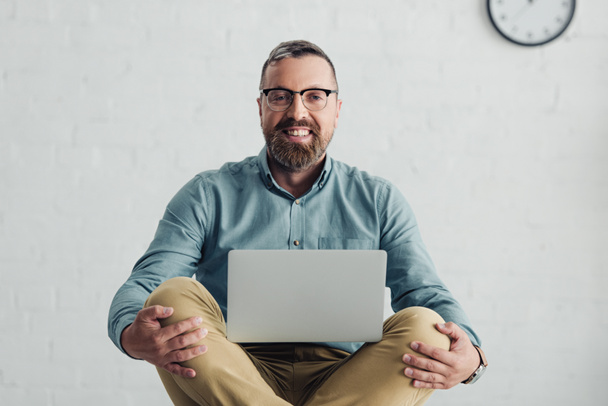 homem de negócios bonito na camisa e óculos sorrindo e segurando laptop
  - Foto, Imagem