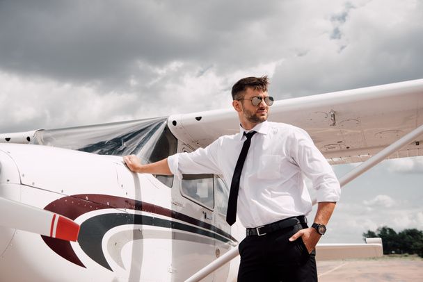 piloto seguro de sí mismo en gafas de sol de pie cerca de avión y mirando hacia otro lado
 - Foto, Imagen