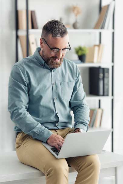 bel homme d'affaires en chemise et lunettes assis sur la table et utilisant un ordinateur portable
  - Photo, image