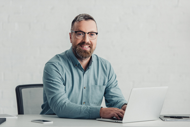 όμορφος επιχειρηματίας σε πουκάμισο και γυαλιά χρησιμοποιώντας φορητό υπολογιστή και κοιτάζοντας την κάμερα  - Φωτογραφία, εικόνα