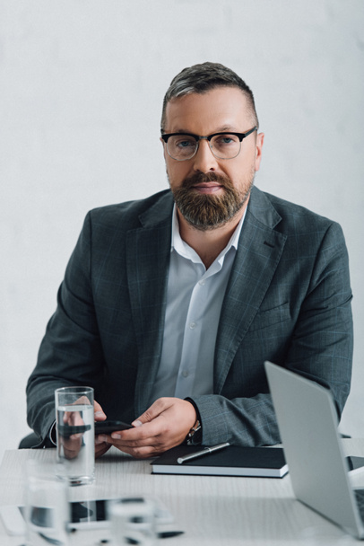 handsome businessman in formal wear and glasses holding smartphone in office  - Fotografie, Obrázek
