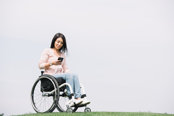 smiling disabled woman in wheelchair using smartphone while resting in park - Foto, Imagen