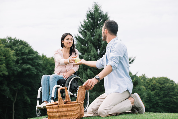 young man giving apple to happy disabled girlfriend while resting together in park - Photo, Image