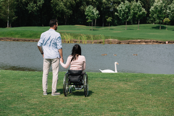 back view of young man holding hands with disabled woman while looking at pond in park - Photo, Image