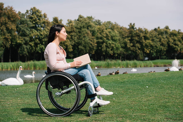 dreamy disabled woman holding book while resting near pond with birds - Photo, Image