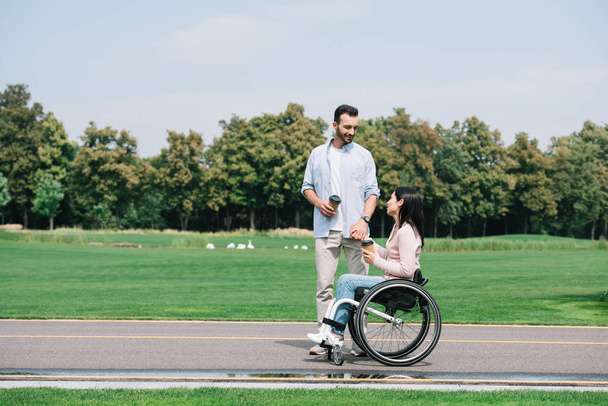 handsome man with disabled girlfriend holding coffee to go while walking in park - Fotoğraf, Görsel