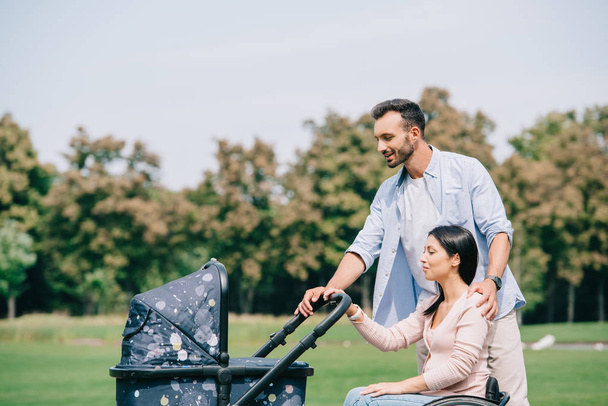 happy man and disabled woman walking with baby carriage in park together - Foto, imagen