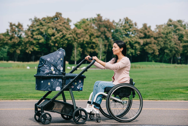 νεαρή γυναίκα με ειδικές ανάγκες σε αναπηρικά αμαξίδια περπατώντας με βρεφική άμαξα στο πάρκο - Φωτογραφία, εικόνα
