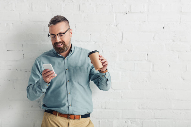 handsome businessman in shirt and glasses holding smartphone and paper cup  - Photo, Image